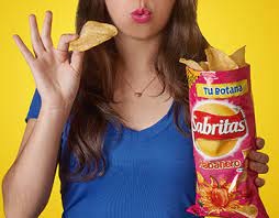 Woman with blue shirt in front of yellow background holding a bag of Sabritas Adobadas with left hand and holding a piece of chip on the right hand
